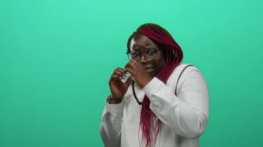 Pembe örgülü ve gözlüklü bir kadın, izole edilmiş yeşil bir arka plan duvarına karşı gergin bir şekilde poz veriyor. Beyaz bir gömlek giyiyor ve modern şehir tarzını canlı bir şekilde ön plana çıkarıyor..