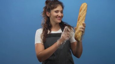 Eldivenli ellerle gülümseyen bir kadın bir baget tutuyor ve mavi stüdyoda onu işaret ediyor; neşeli taze ekmek.