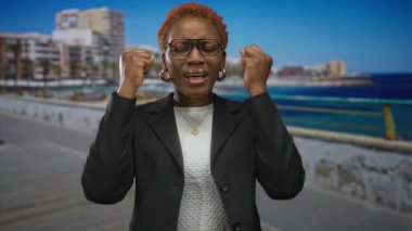 Siyah ceketli ve gözlüklü kadın deniz kenarında ağzı açık bir şekilde şehir rıhtımındaki binaların yanında kol kola yürür..