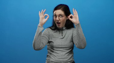 Esmer saçlı bir kadın, gri bir süveter ve gözlüklü, parlak mavi bir arka plana karşı güvenle gülümseyen hoş bir jest yapıyor..