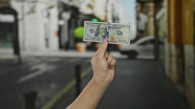 Adam bizi bir şehir caddesinde tutuyor, açık bir şehir ortamında yüz dolarlık banknotlar sergiliyor, finans ve ekonomik değişimin özünü yakalıyor..