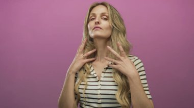 Sarışın bir kadın zarif bir şekilde pembe arka plan duvarına karşı poz veriyor, çizgili gömleğinin içinde zarafet ve sakinlik yayıyor, sakin ve şık bir görsel izlenim yaratıyor..