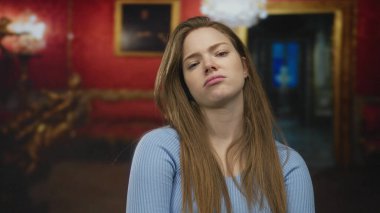 Kollarını kavuşturmuş ve dudaklarını bükmüş bir kadın sanat galerisinde çerçeveli resimler ve avizeyle duruyor..