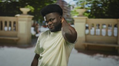 Bir adam taşın yanında boynuna sağ eliyle dokunuyor, açık tişört giyiyor ve hafif bir gülümseme takınıyor; şakacı benlik bilinci.