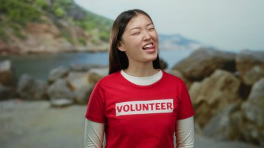 Deniz kenarında gülümseyen gönüllü tişörtlü bir kadın. Kayalık arka planda neşeli bir açık hava enerjisi ve canlı bir toplum ruhu sergiliyor..