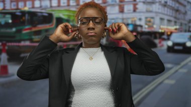 Gözlüklü ve spor ceketli kadın, yoğun şehir trafiğinde kulaklarını kapatıyor; duyusal aşırı yüklenme.