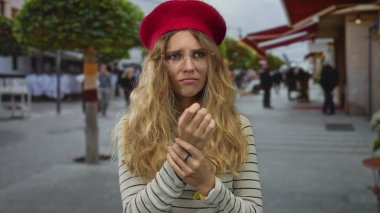 Sarışın, genç bir bere takmış, gözlüklü, çizgili bluzu olan bir kadın sokakta bileğini kavrıyor ve izliyor..