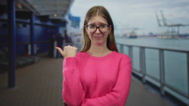 Pembe kazaklı genç kadın, deniz manzaralı limana bakan güvertenin arkasını işaret ediyor..