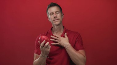 Stüdyoda eli göğsünde kırmızı elma tutan adam kırmızı arka planda; düşüncelere dalma samimiyeti.