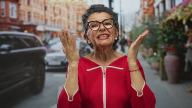 Gri saçlı, gözlüklü ve kırmızı elbiseli yaşlı bir kadın, arka planda arabalar ve binalar olan bir şehir caddesinde hareketli şehir atmosferini yakalıyor..
