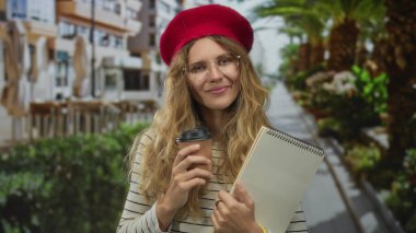 Sarışın bereli, gözlüklü ve şehir ortamında bir kafede defter tutan bir kadın elinde kahveyle sokakta gülümsüyor..