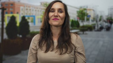 Şehir caddesinde güvenle gülümseyen bir kadın, canlı ruj ve günlük kıyafetlerle elini uzatarak, dost canlısı, kentsel bir atmosfer yaratıyor..