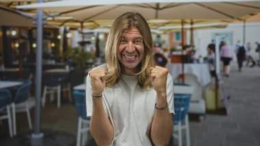 Beyaz tişörtlü, uzun sarı saçlı bir adam, açık hava restoranlarının terasında şemsiyelerin altında yumruklarını sıkıyor; heyecan verici zafer kutlamaları zaferi.