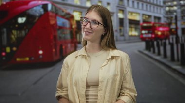 Yoğun bir şehir caddesinde kırmızı bir çift katlı otobüsün yanında gülümseyen gözlüklü genç sarışın kadın, iyimserlik..
