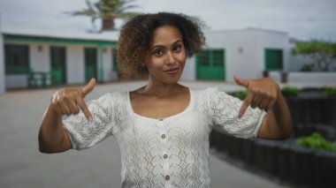 Kadın her iki parmağını da bir binanın avlusunun önüne işaret ediyor, beyaz örgü bir bluz giyiyor ve kameraya gülümsüyor; kendine güven.