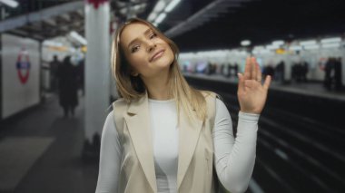 Kapalı tren istasyonundaki platforma gülümseyen, bulanık insanlarla çevrili bir kadın ve tren yolu işaretleri günlük seyahat enerjisini yansıtıyor..