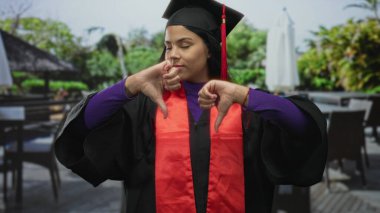 Mezuniyet elbisesi ve kepi giymiş bir kadın havuz kenarındaki bir restoranın terasında baş parmağıyla jest yapıyor..