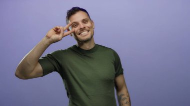 İki eliyle barış işareti yapan çekici genç bir adam canlı mor bir arka plana karşı neşeyle gülümsüyor. Dövmelerini ve günlük tarzını bir stüdyo ortamında sergiliyor.