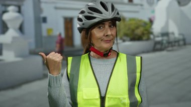 Miğferli ve yüksek görüşlü bir yelek giyen kıdemli İspanyol kadın şehir sokaklarında el kol hareketi yaparak şehir ortamında güvenlik ve farkındalık sağlıyor..