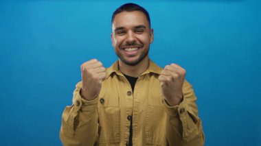 Erkek, canlı mavi stüdyo ortamında geniş bir gülümseme ile yumruklarını sırıtır ve sıkar; zafer enerji tutkusunu kutlar.