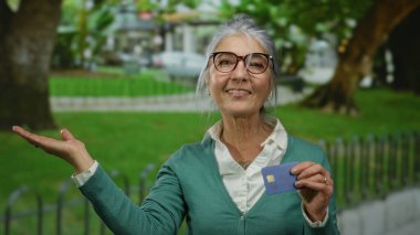 Parktaki son sınıf öğrencisi kadın elinde kredi kartıyla güneşli bir günde açık havada neşe saçıyor, gözlük takıyor ve yeşil hırka giyiyor..