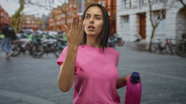 Kadın, bisiklet ve tuğla binalı bir caddede pembe deterjan şişesini tutarken burnunu çimdikliyor; koku iğrenmeyle ilgili..