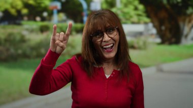 Kırmızı gömlekli yaşlı bir kadın, canlı bir parkta gülümserken, taştan jest yapıyor. Yeşillik ve görünür yürüyüş yolu var, neşe ve coşku saçıyor..