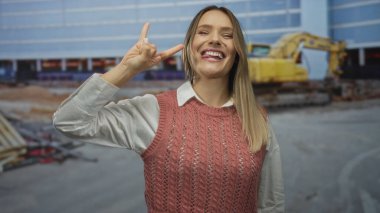 Dışarıdaki inşaat alanında kazıcıyla gülümseyen, kaya jestleri yapan, pembe kazak giyen, bina ortamında heyecan ve neşeyi simgeleyen bir kadın..