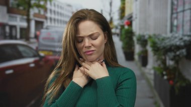 Çiçek saksısı ve park halindeki arabalarla dolu bir caddede boğazını sıkan genç bir kadın..