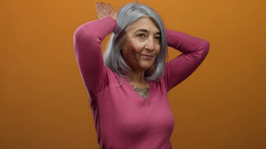 Gri saçlı yaşlı kadın, elleri turuncu arka planda, pembe bir kazak giyiyor, mizahi ve neşeli bir sahne yaratıyor..