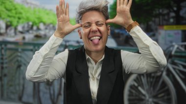 Beyaz bir kadın, şehrin bir caddesindeki bir kanal köprüsünde park halindeki bir bisikletin yanında el sallıyor..