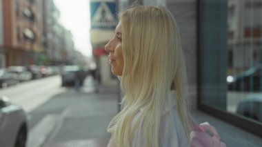 Park halindeki arabaların ve vitrinlerin yanında pembe sırt çantalı ve uzun sarı saçlı bir kadın yürüyor..