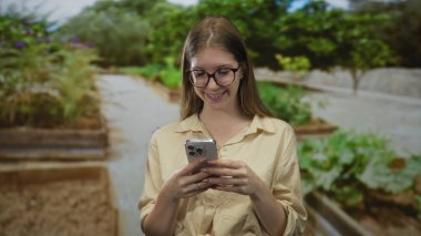 Gözlüklü ve bej gömlekli kadın orman yolunda akıllı telefonu yüze yakın tutuyor; dijital bağlantı memnuniyeti.