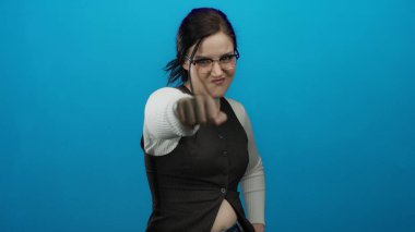 Yumruğunu kameraya doğru uzatan, parlak mavi arka plana karşı kararlı bir ifadeye sahip, gözlük takan ve günlük giyinen, güçlü, kendine güvenen, odaklanmış, güzel bir kadın.