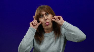 Katı mavi bir arka plana karşı komik suratlar yapan bir kadın, ellerinin yanaklarında olduğu mizah ve ifadeyi vurgulayan, tecrit edilmiş bir ortamda son sınıf İspanyol kadınını gösteren bir kadın..