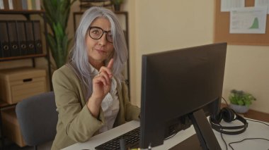 Gri saçlı, gözlüklü son sınıf öğrencisi bir kadın ofiste bilgisayarın başında oturuyor. Kapalı bir odada parmağı olan bir jest yapıyor..