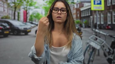 Çimdiklenmiş parmakları olan İspanyol genç bir kadın gözlük ve kot gömlek giyerek şehir caddesinde kameraya doğru hareket ediyor..