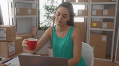 Dizüstü bilgisayarındaki genç İspanyol kadın, kırmızı bardağı binadaki istiflenmiş paketlerin yanında sessizce tutuyor..