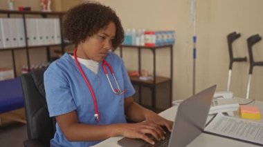 Mavi önlük giyen kadın doktor ve klinikteki masamda dizüstü bilgisayarda yazan kırmızı steteskop; şefkat gösterme uzmanlığı.