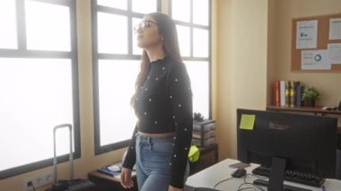 Modern ofis ortamında kendine güvenen, gözlüklü, uzun saçlı, profesyonelliği somutlaştıran ve doğal ışık alan bir iç mekan ortamında odaklanan bir kadın..