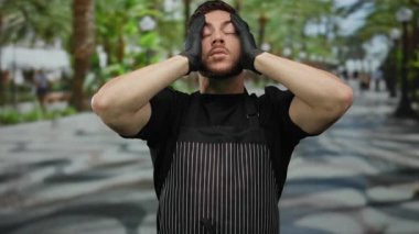 Sakallı ve önlüklü genç adam şehir sokaklarında eldiven takıyor. Dalgın görünüyor..