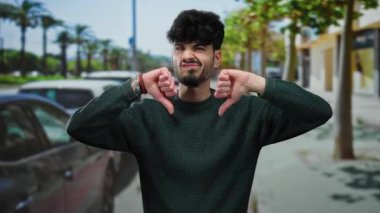 Young man with a beard and curly hair gives a double thumbs-down on an urban street lined with cars and trees, expressing disapproval in a city setting.