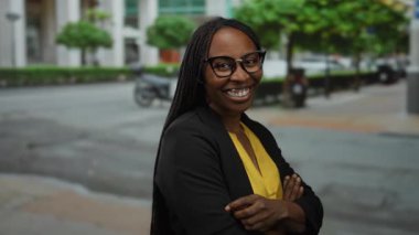 Şehir caddesinde güvenle gülümseyen bir kadın gözlük takıyor ve arka planda ağaçlar ve binalar olan sarı bir bluz giyiyor..