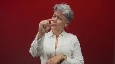 Kadın, beyaz gömlek ve tahta kol saati takarken stüdyoda kırmızı arka plana karşı küçük boy göstermek için parmaklarını çimdikliyor; şüphecilik.