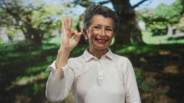Gri saçlı, gülümseyen yaşlı kadın yeşil ağaçlar ve yemyeşil bitkilerle çevrili güneşli bir parkta el hareketi yapıyor.