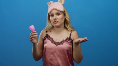 Gece elbisesi giymiş genç bir kadın elinde mavi arka planda karışık bir ifadeyle bir bardak tutuyor.