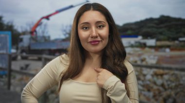 Genç İspanyol kadın açık hava inşaat sahasında vinç ve molozların önünde parmak göğse işaret ediyor..