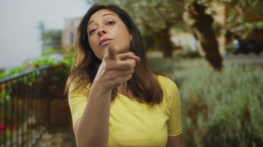 Sarı gömlekli kadın yeşil yeşilliklerin ortasında bina cephesinde parmağını sağa doğru gösteriyor; iddia makamı.