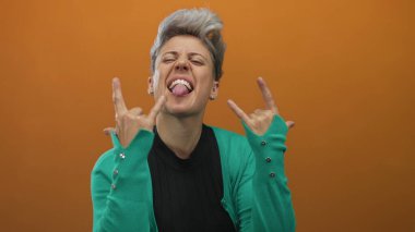 Kısa gümüş saçlı kadın, turuncu stüdyo ortamında taş boynuzlarını ve dilini gösteriyor;.