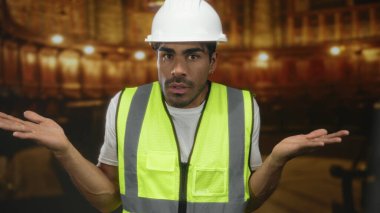 Kask ve güvenlik yeleği giyen İspanyol genç adam kilise binasında omuz silkiyor; belirsizlik.